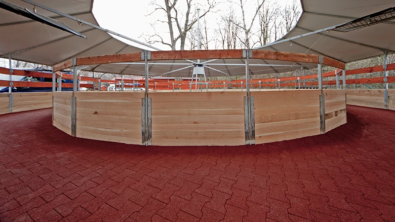 Zdjęcie przedstawia nowoczesną, zadaszoną karuzelę dla koni. Tor wyłożony jest czerwonym brukiem z granulatu gumowego – antypoślizgowym, przepuszczalnym i przyjaznym dla stawów. Elastyczna nawierzchnia zapewnia bezpieczny ruch koni w każdych warunkach pogodowych i chodach. Utwardzona ścieżka jest trwała, stabilna i łatwa w utrzymaniu. Obiekt nadaje się idealnie do ośrodków jeździeckich, stadnin i klinik weterynaryjnych, które stawiają na komfort, trwałość i niskie koszty eksploatacji.