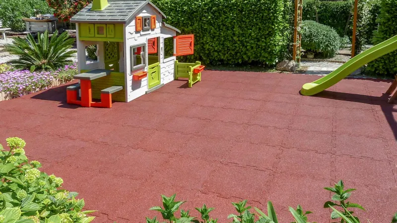 Plac zabaw w ogrodzie to marzenie wielu dzieci. Na powierzchni z czerwonych bezpiecznych płyt amortyzujących WARCO zainstalowano plastikowy domek do zabawy.