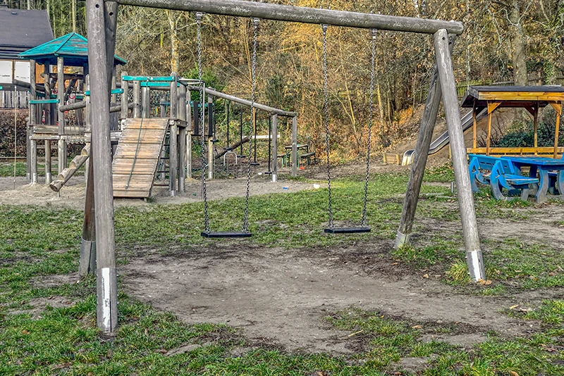 Trawiaste podłoże nie jest odpowiednie jako zabezpieczenie przed upadkiem na publicznym placu zabaw. Trawa pod huśtawką jest uszkodzona i tworzy się błoto.
