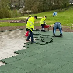 Grupa pracowników układa zielone płyty WARCO do gry w piłkę w deszczu na betonowym obszarze wyłożonym płytkami.