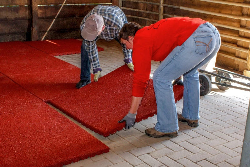 Dwóch mężczyzn układa czerwone, wodoprzepuszczalne, antypoślizgowe maty do stajni WARCO w boksie dla koni bezpośrednio na betonowej kostce brukowej.