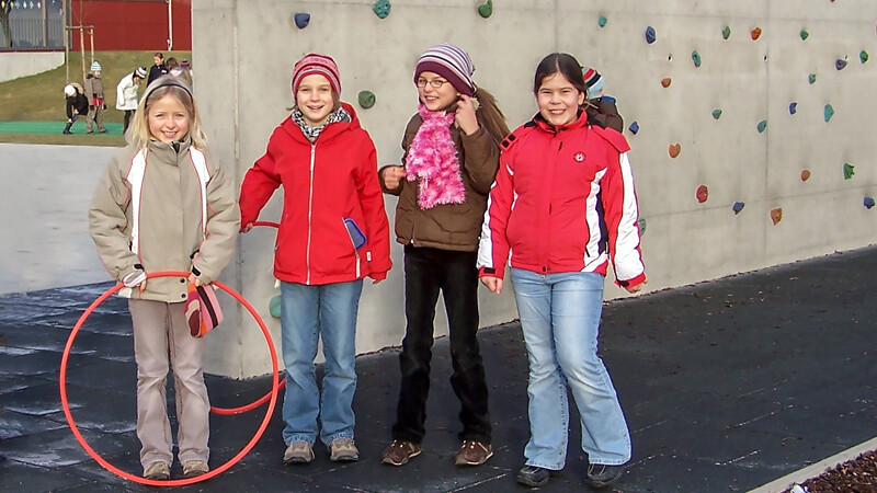 Cztery uśmiechnięte dziewczynki stoją przed betonową ścianką wspinaczkową na placu zabaw z płytami amortyzującymi WARCO chroniącymi w razie upadku z wysokości.