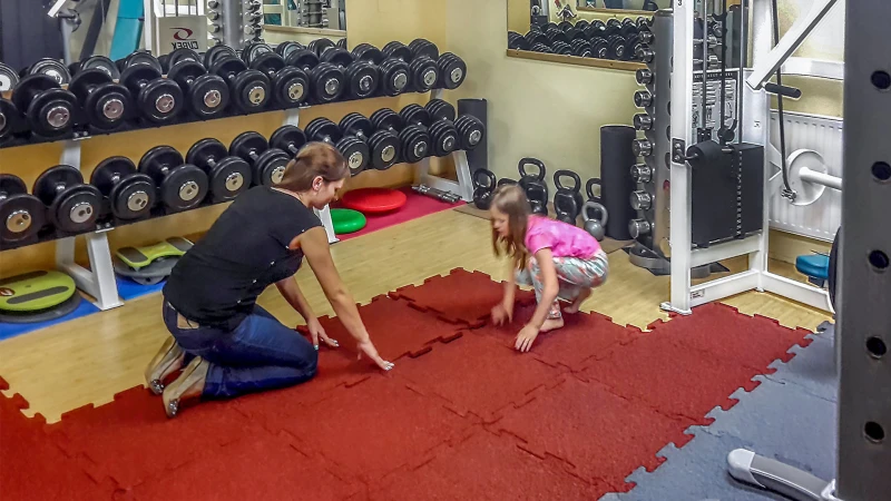 Mama z córką układają płyty sportowe WARCO z łączeniem na zamkiem bezpośrednio na panelach w domowej siłowni wyposażonej w sprzęt do ćwiczeń.