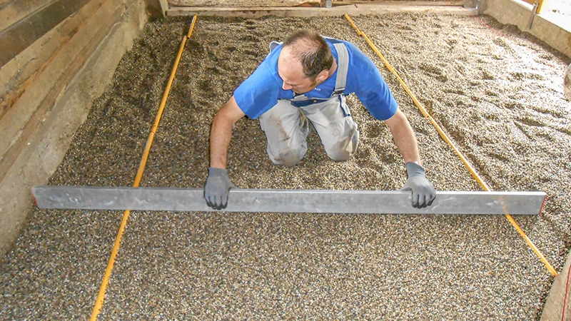 Układanie nowej nawierzchni w stajni: mężczyzna wyrównuje podsypkę żwirową za pomocą łaty murarskiej. Żwir służy jako podkład pod podłoże boksowe z granulatu gumowego. Powierzchnia ograniczona jest po bokach drewnianymi lub betonowymi ścianami. Prace wykonywane są starannie i precyzyjnie – typowe dla prawidłowego wykonania podłoża przyjaznego koniom.