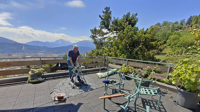 Duży balkon z szarymi płytkami balkonowymi WARCO i widokiem na góry. Mężczyzna pracuje przy układaniu płyt.