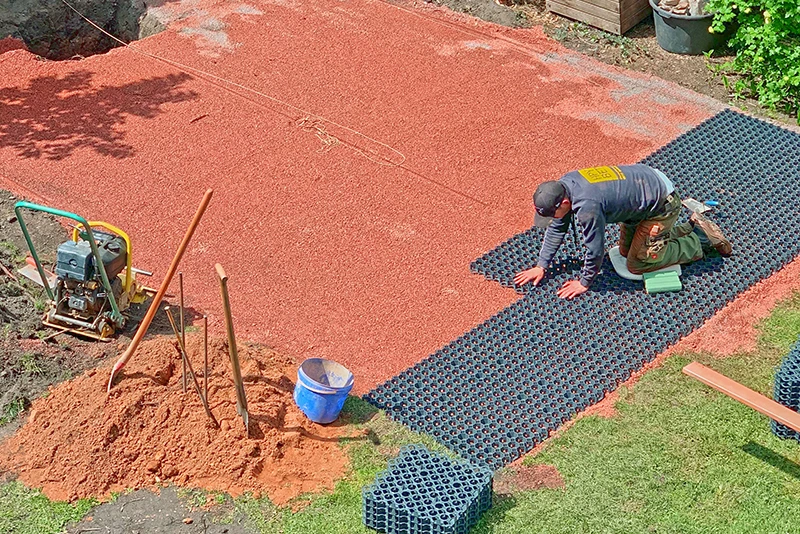 Mężczyzna układa zielone płyty w kształcie puzzli od WARCO na plastikowych kratkach wypełnionych żwirem.