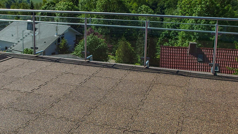 Na zdjęciu widoczny jest duży balkon z przezroczystą balustradą, z którego rozciąga się widok na okoliczne dachy i bujną zieleń. Podłoże wyłożono płytami puzzle WARCO z granulatu gumowego w ciepłym, brązowym odcieniu. Dzięki zazębiającym się krawędziom powstaje jednolita powierzchnia, która jest antypoślizgowa i odporna na warunki atmosferyczne—doskonała do zastosowań zewnętrznych. Dodatkowo nawierzchnia amortyzuje uderzenia i zapewnia dobrą izolację akustyczną. Łatwa pielęgnacja sprawia, że to trwałe i wygodne rozwiązanie na balkony i tarasy.
