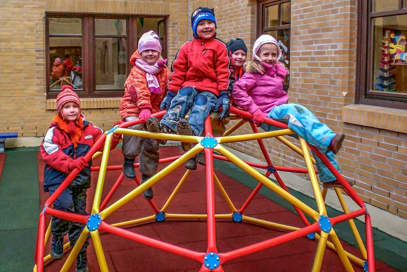 Uśmiechnięte dzieci siedzą na konstrukcji wspinaczkowej na placu zabaw z czerwonymi i zielonymi płytami amortyzującymi WARCO, które zapewniają ochronę przed poważnymi urazami w przypadku upadku w wysokości.